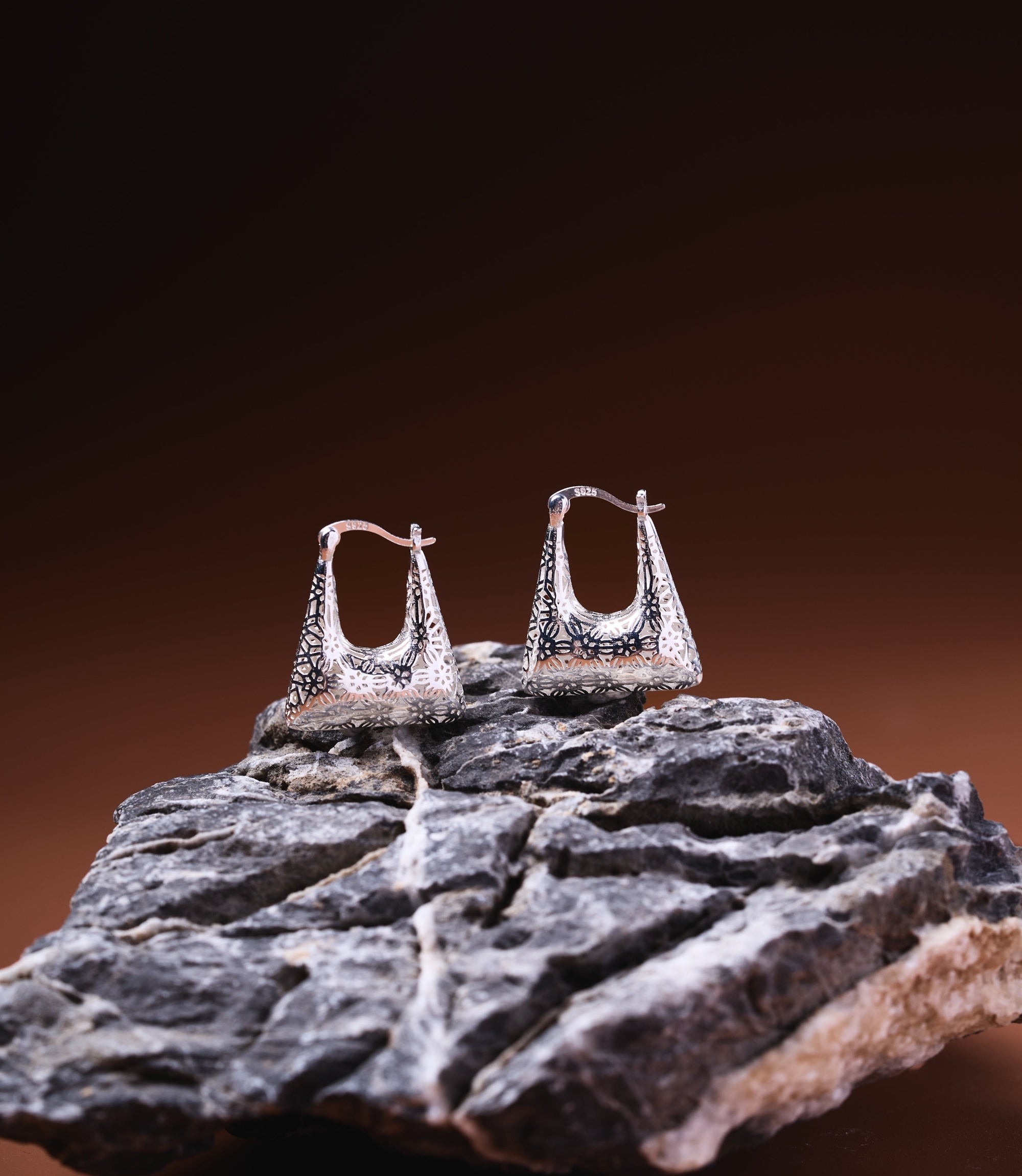 Naya Sterling Silver 925 hoop earrings with trapezoid shape and oxidised floral engravings, displayed on dark stone